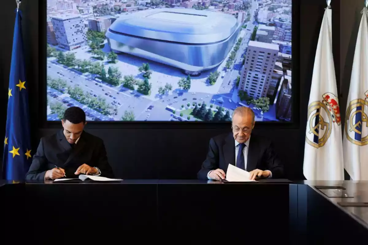Dos hombres sentados firmando documentos en una mesa, con banderas oficiales a los lados y una imagen de un estadio moderno proyectada en la pantalla detrás de ellos Dos hombres sentados firmando documentos en una mesa, con banderas oficiales a los lados y una imagen de un estadio moderno proyectada en la pantalla detrás de ellos