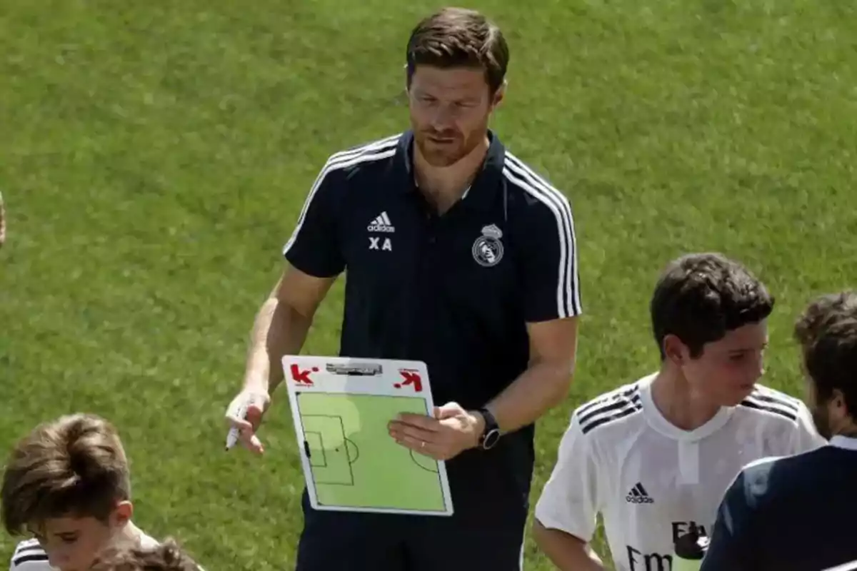 Un entrenador de fútbol con uniforme del Real Madrid sostiene una pizarra táctica mientras habla con jóvenes jugadores en un campo de césped. Un entrenador de fútbol con uniforme del Real Madrid sostiene una pizarra táctica mientras habla con jóvenes jugadores en un campo de césped.
