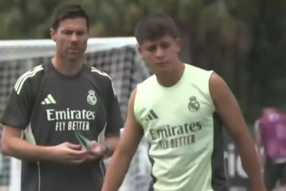 Two people wearing Real Madrid training gear are on a soccer field; one of them appears to be a coach and the other a player.