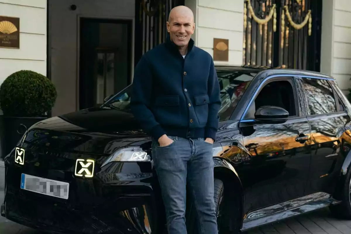 A bald man in a dark blue jacket and jeans is standing in front of a black car parked in front of an elegant building.