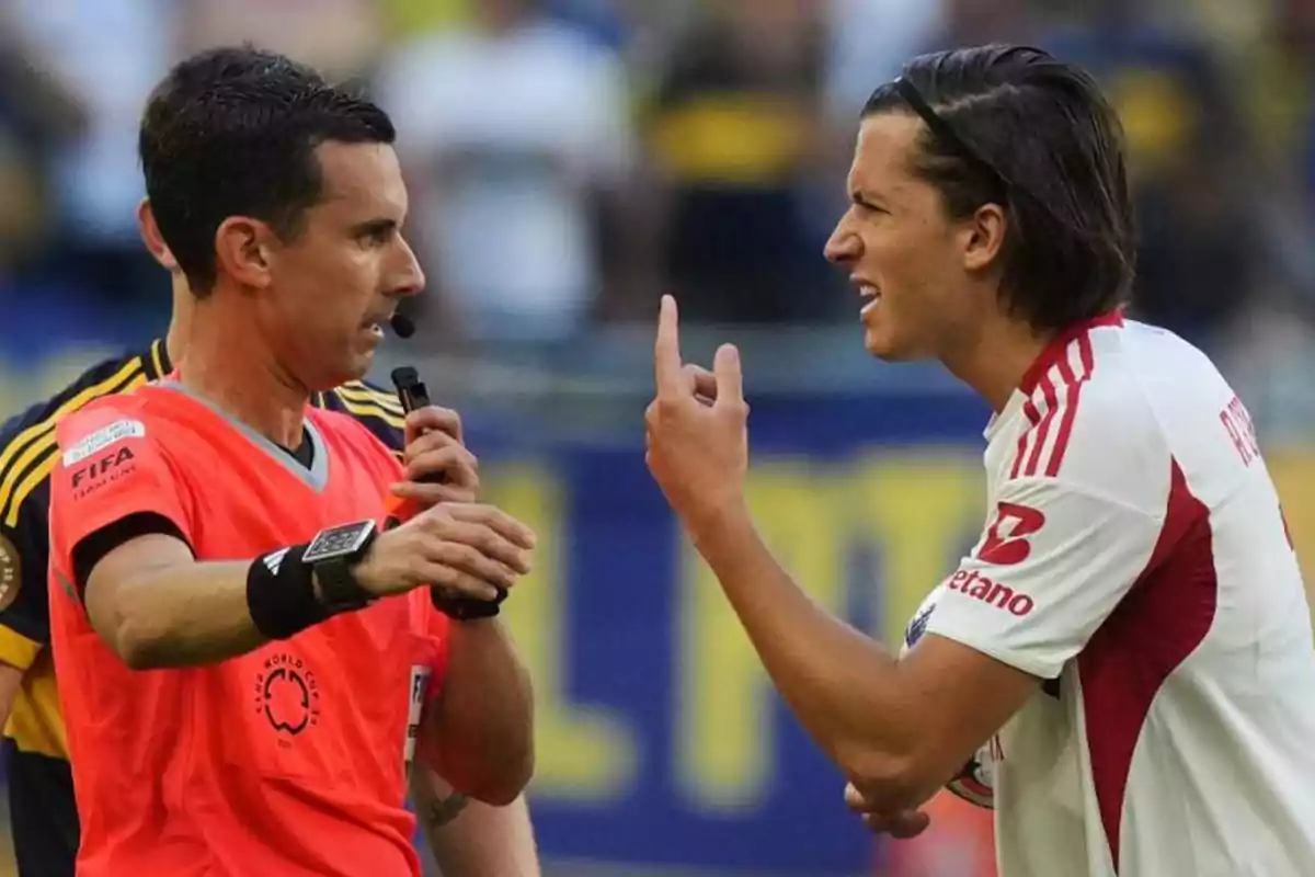Un árbitro de fútbol sostiene un silbato mientras un jugador con uniforme blanco y rojo le reclama gesticulando con la mano durante un partido Un árbitro de fútbol sostiene un silbato mientras un jugador con uniforme blanco y rojo le reclama gesticulando con la mano durante un partido