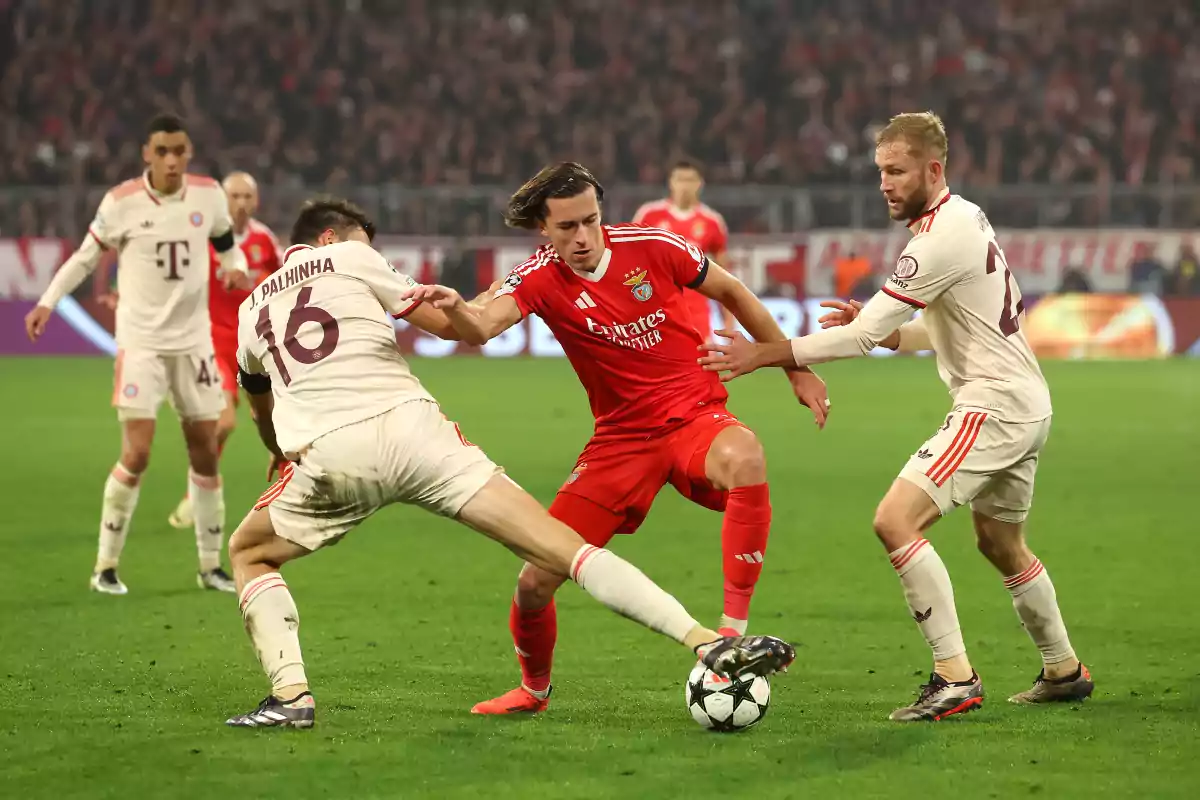A player in a red uniform tries to advance with the ball while being marked by two opposing team players dressed in white in a stadium full of spectators. A player in a red uniform tries to advance with the ball while being marked by two opposing team players dressed in white in a stadium full of spectators.
