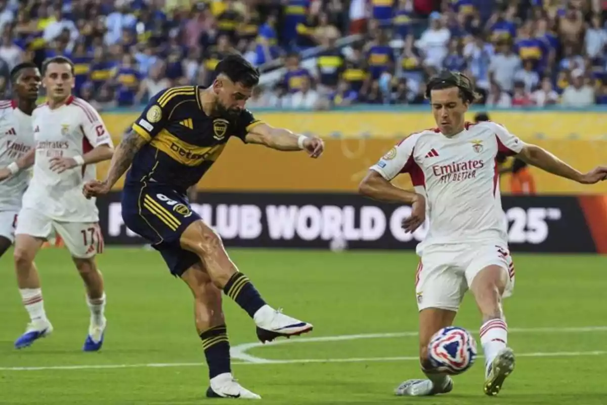 A Boca Juniors player kicks the ball while a Benfica defender tries to block the shot during a soccer match with spectators in the stands.