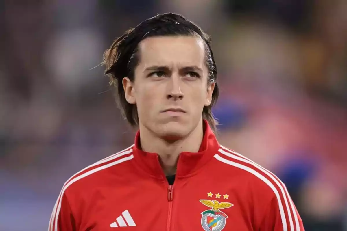 A player in a red jacket with the Benfica logo, looking forward with a serious expression. A player in a red jacket with the Benfica logo, looking forward with a serious expression.