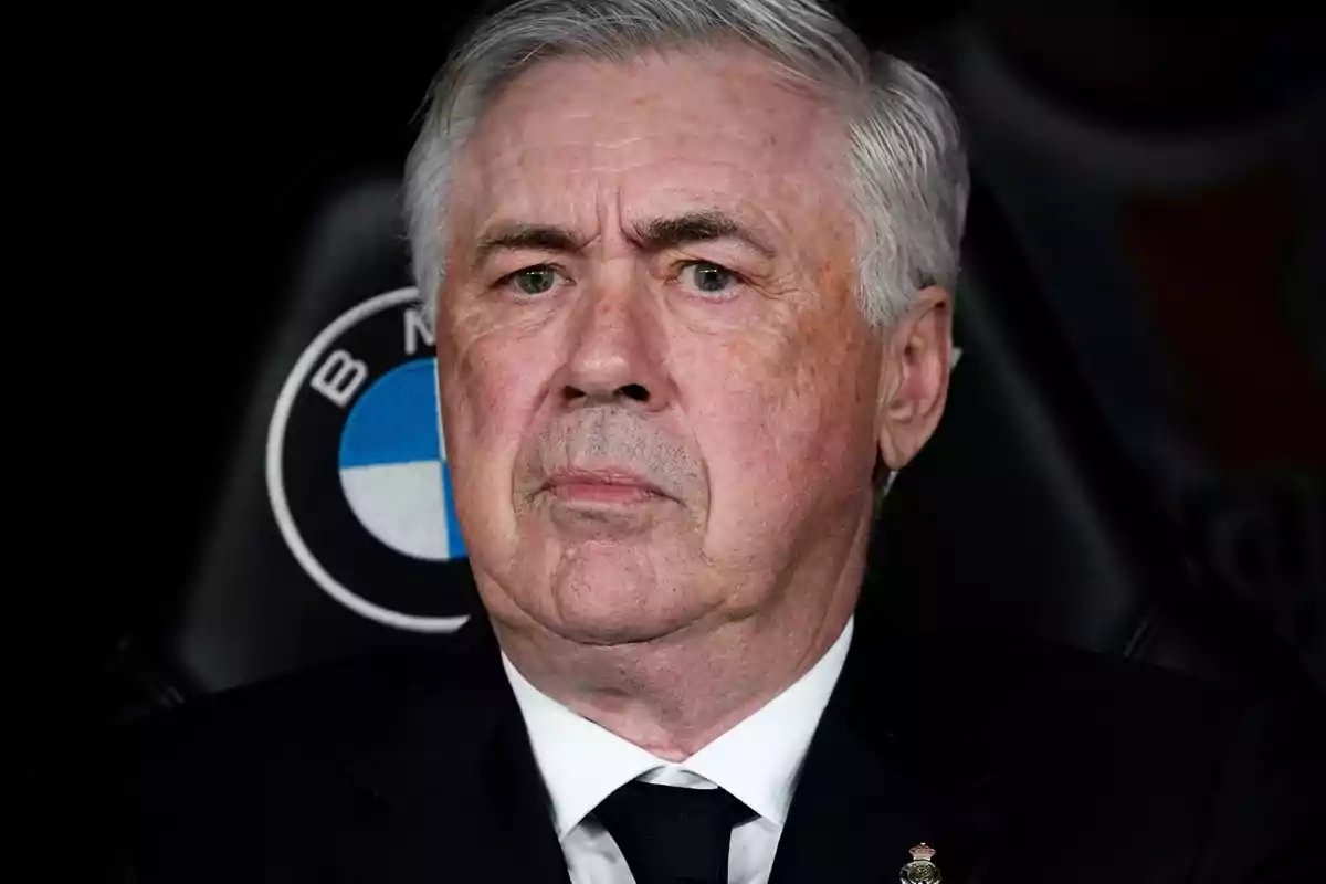 A gray-haired man with a serious expression is sitting in a seat with a visible logo in the background. A gray-haired man with a serious expression is sitting in a seat with a visible logo in the background.