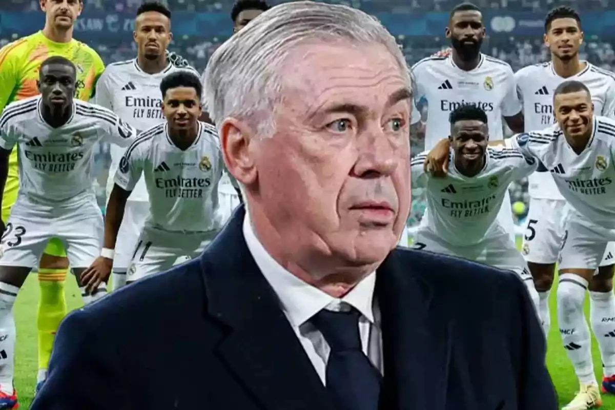 Un entrenador de fútbol con traje oscuro está en primer plano, mientras que detrás de él se encuentra un equipo de jugadores con uniformes blancos posando en un campo de fútbol.