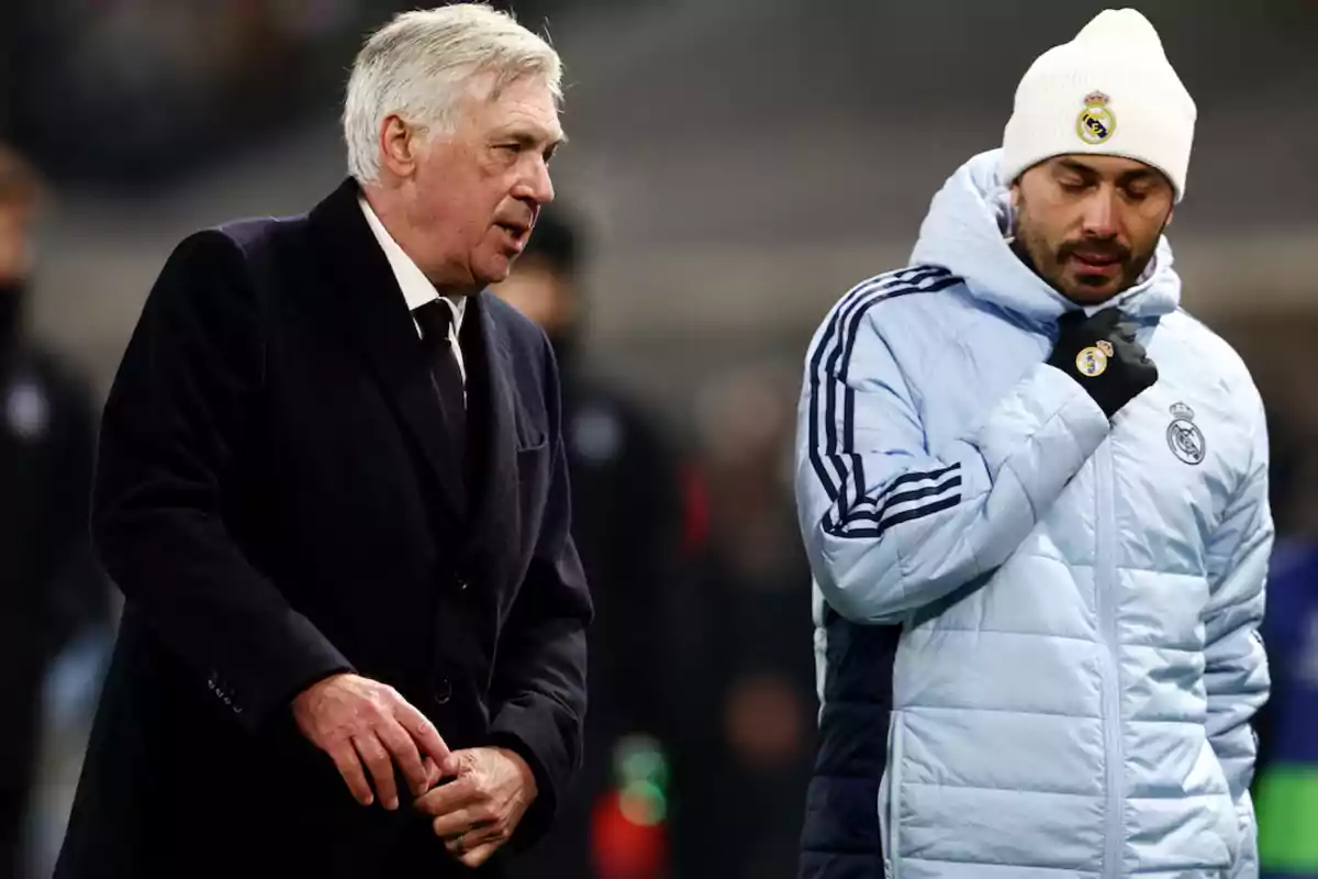 Dos hombres con ropa de invierno del Real Madrid conversan en un campo de fútbol.