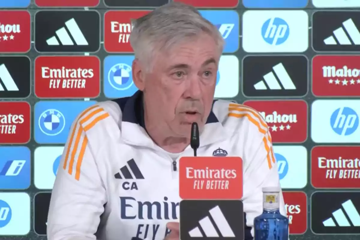 A man in a white sports jacket with orange details speaks at a press conference with a background of sponsor logos. A man in a white sports jacket with orange details speaks at a press conference with a background of sponsor logos.