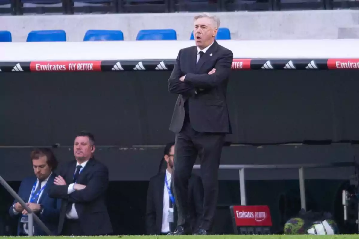 A man in a black suit with his arms crossed in the technical area of a soccer stadium. A man in a black suit with his arms crossed in the technical area of a soccer stadium.