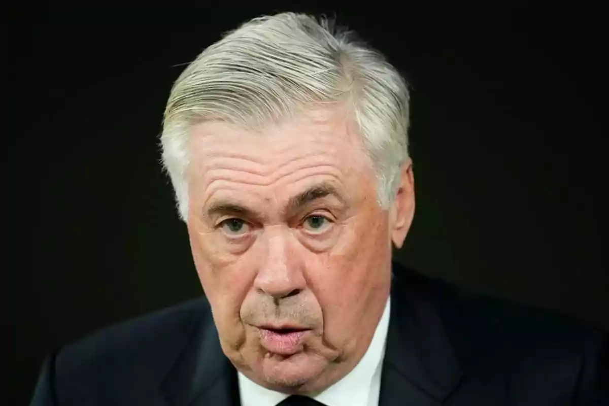 A gray-haired man with a serious expression wears a dark suit against a black background.