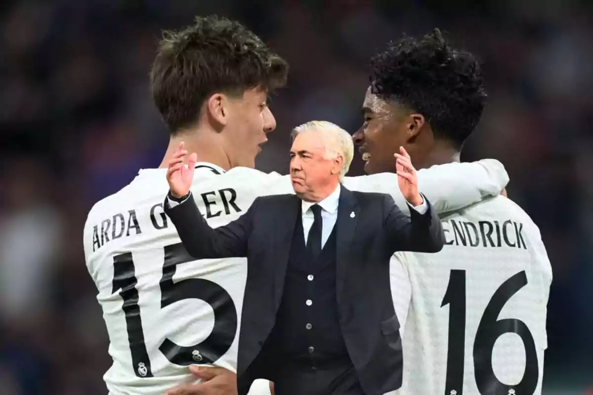 A soccer coach in a black suit raises his arms while two players in white jerseys hug in the background.