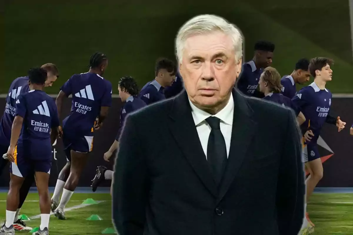 A man in a suit is in the foreground while a group of players trains on the field behind him.