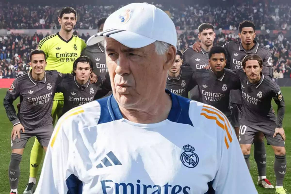 Un entrenador de fútbol con gorra blanca y camiseta del Real Madrid está en primer plano mientras un grupo de jugadores del equipo posa detrás de él en un estadio lleno de espectadores.