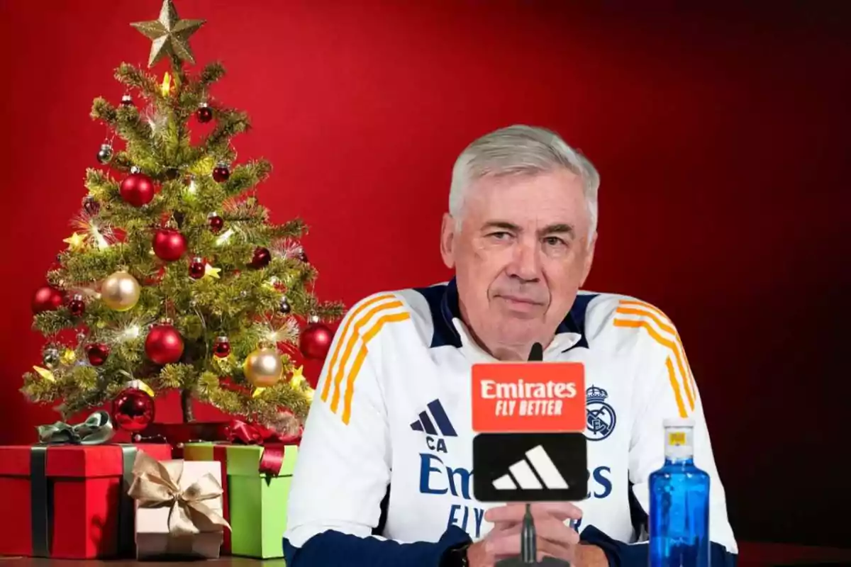 Un hombre con uniforme deportivo del Real Madrid está sentado frente a un micrófono con un fondo rojo y un árbol de Navidad decorado con luces y esferas.