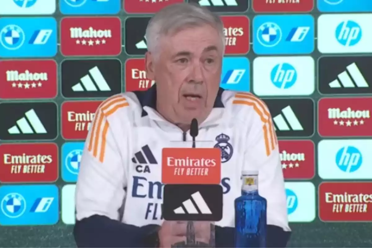 A man in a white and orange Real Madrid sports uniform is speaking at a press conference, with a microphone in front of him and a backdrop of sponsor logos.