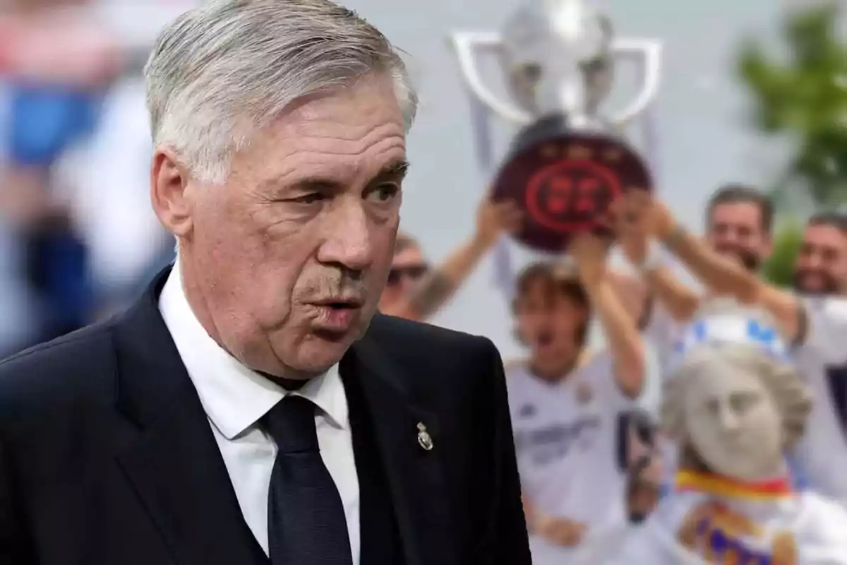 A gray-haired man in a dark suit in the foreground with people celebrating and raising a trophy in the background.