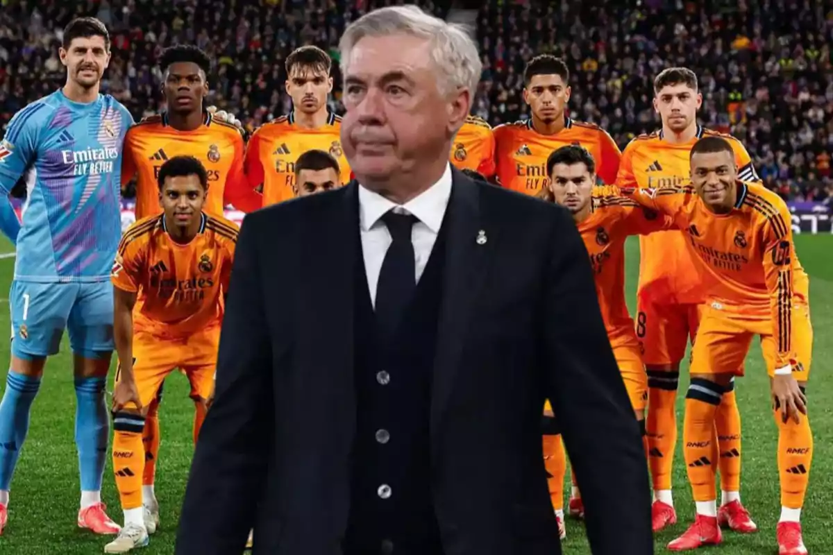 A soccer coach in a dark suit stands in front of a group of players in orange uniforms on a soccer field.