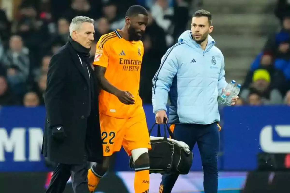 Un jugador de fútbol del Real Madrid, vestido con un uniforme naranja, camina acompañado por dos personas en un estadio.