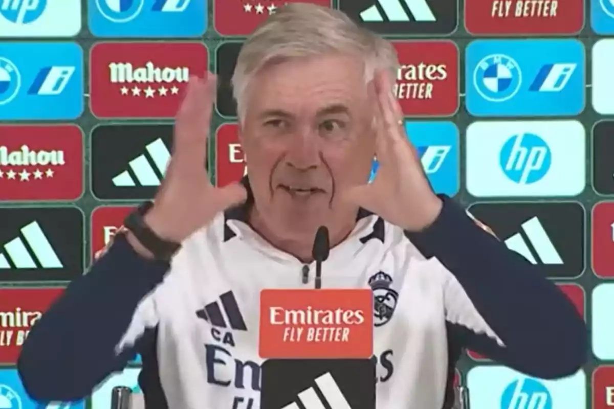 A man at a press conference gestures with his hands as he speaks, against a background of sponsor logos.