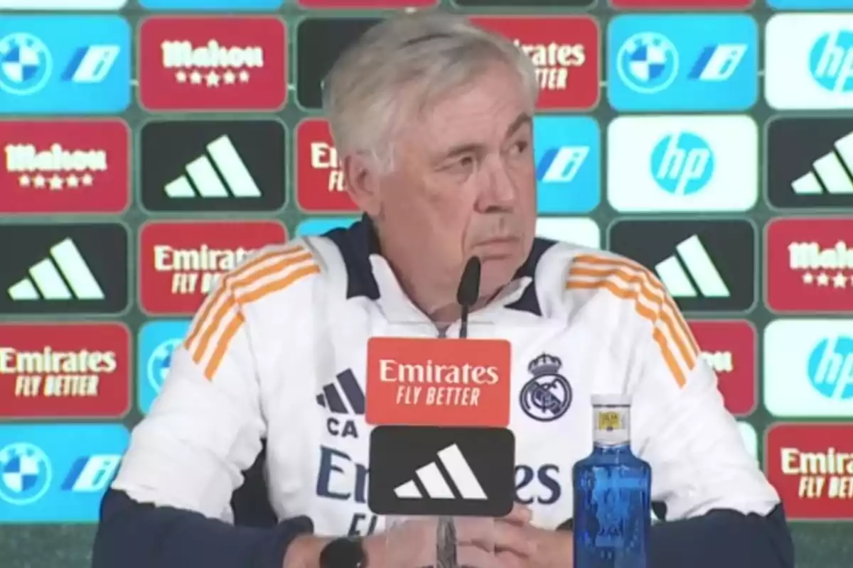 A man at a press conference with a microphone in front of him and a background with sponsor logos.