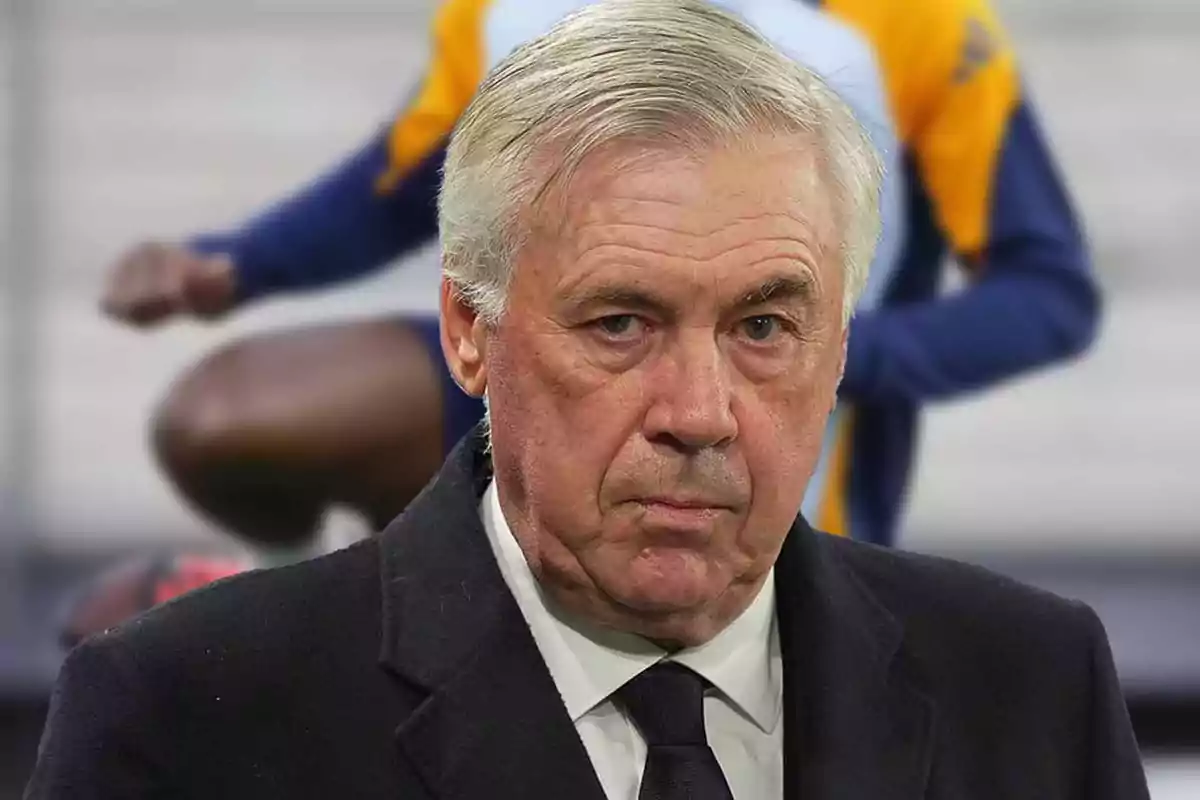 A man with gray hair and a serious expression wears a dark coat with a tie in a sports environment.