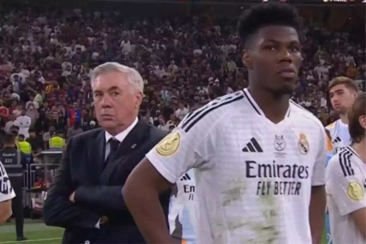 Un entrenador de fútbol con traje oscuro observa el campo mientras un jugador con uniforme blanco está de pie cerca de él en un estadio lleno de espectadores.