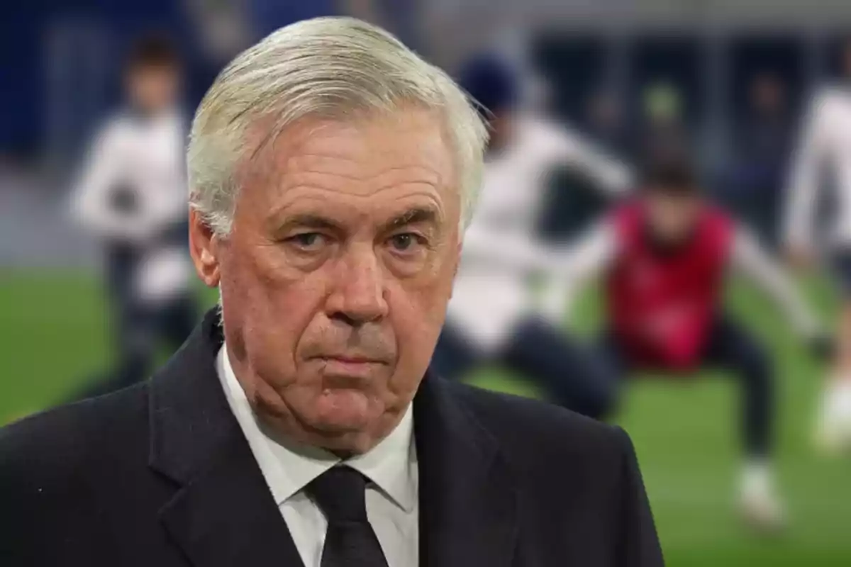 A man with gray hair and a serious expression with a blurred background of people training on a soccer field.