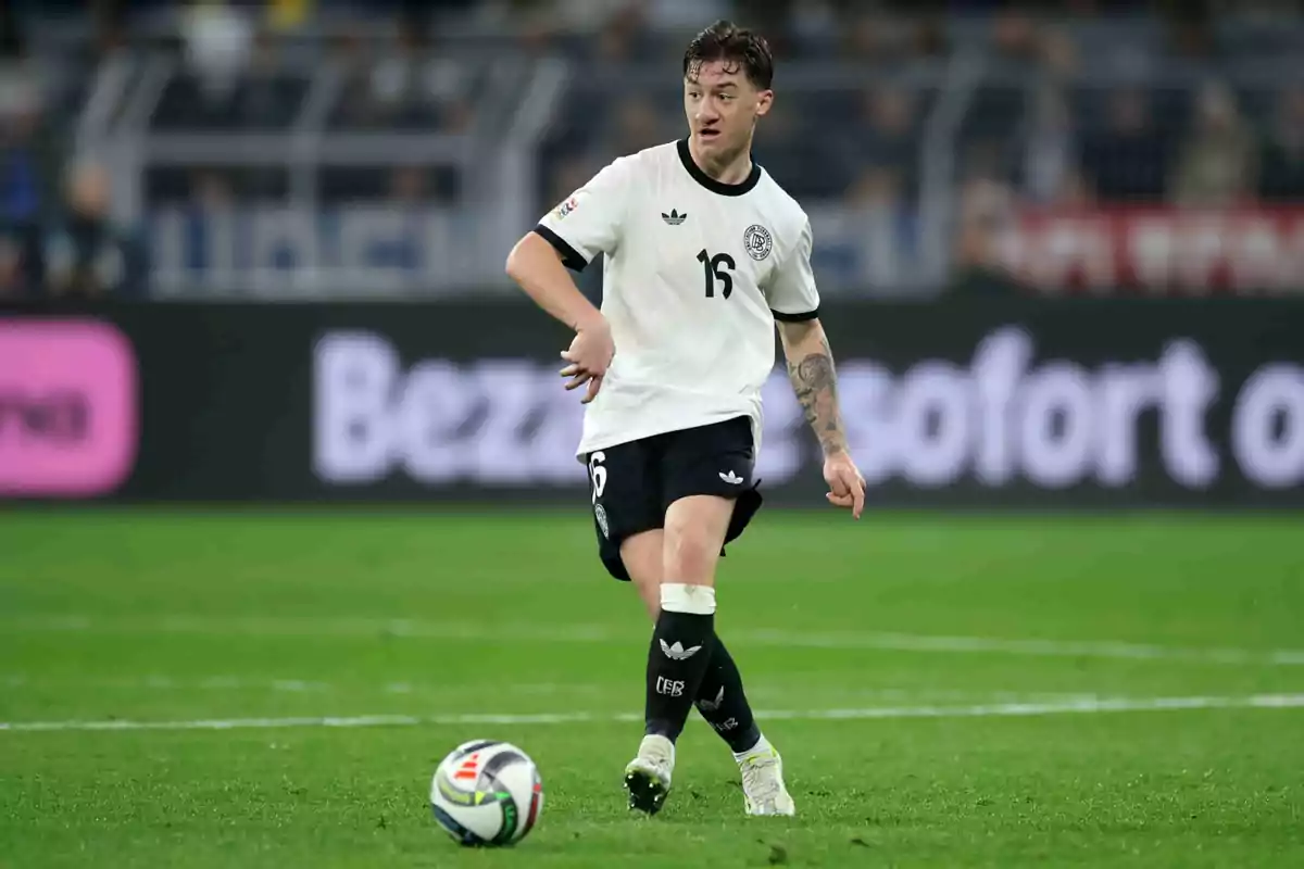 Un jugador de fútbol con el número 16 en su camiseta blanca y pantalones cortos negros está en el campo durante un partido, con un balón cerca de sus pies. Un jugador de fútbol con el número 16 en su camiseta blanca y pantalones cortos negros está en el campo durante un partido, con un balón cerca de sus pies.