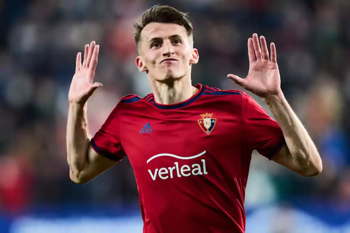 Un jugador de fútbol con uniforme rojo levanta las manos en un gesto de celebración en un estadio. Un jugador de fútbol con uniforme rojo levanta las manos en un gesto de celebración en un estadio.
