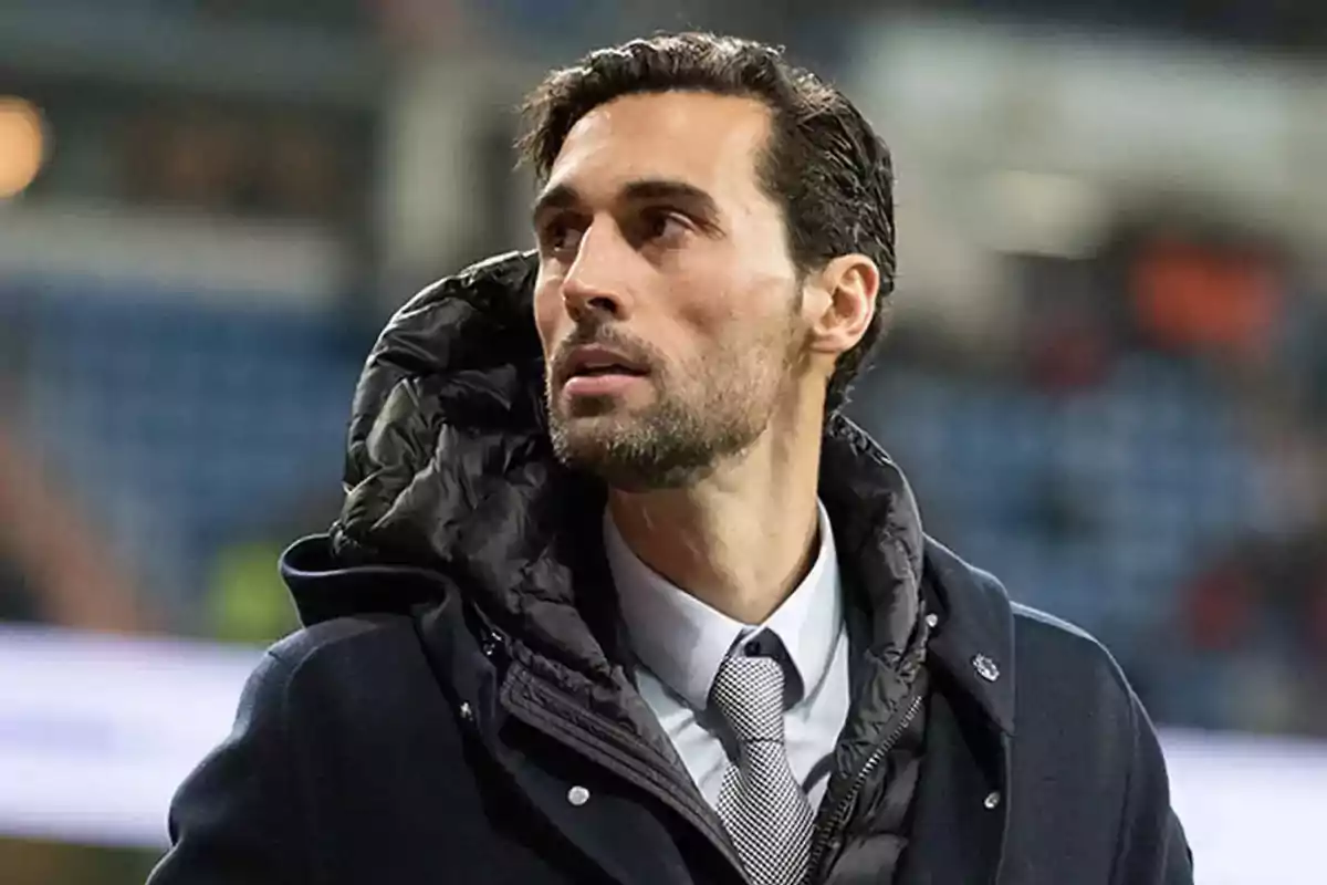Un hombre con barba y cabello oscuro lleva un abrigo y una corbata mientras mira hacia un lado en un entorno urbano. Un hombre con barba y cabello oscuro lleva un abrigo y una corbata mientras mira hacia un lado en un entorno urbano.