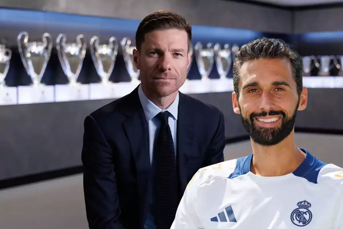 Two men pose in front of a display case filled with Champions League trophies; one of them is wearing a suit and the other is wearing a Real Madrid jersey.