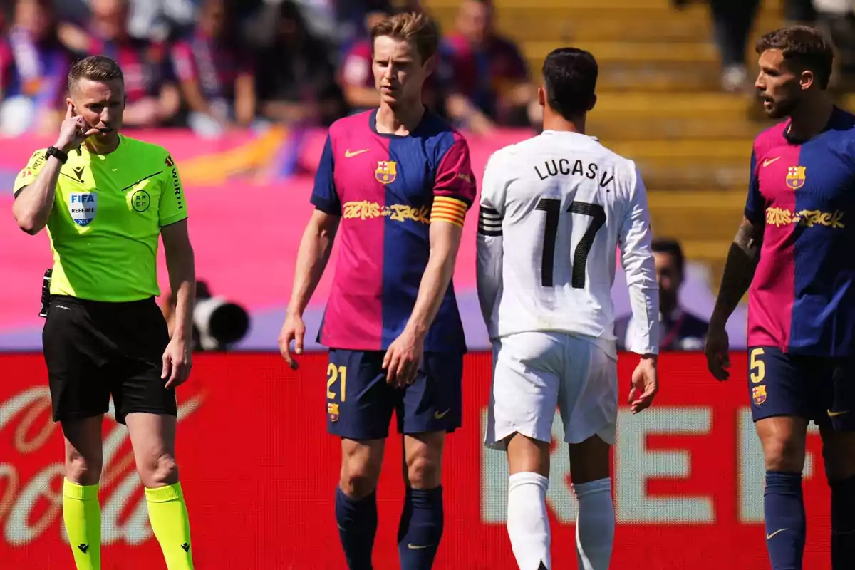Un árbitro de fútbol se comunica por radio mientras dos jugadores del equipo con uniforme azul y rojo y uno del equipo con uniforme blanco están en el campo durante un partido.