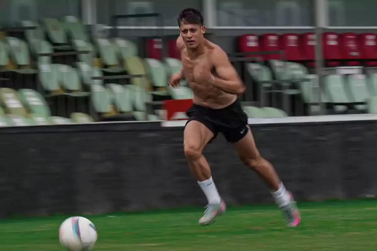 Joven sin camiseta corriendo en una cancha de fútbol mientras persigue un balón con gradas vacías al fondo Joven sin camiseta corriendo en una cancha de fútbol mientras persigue un balón con gradas vacías al fondo