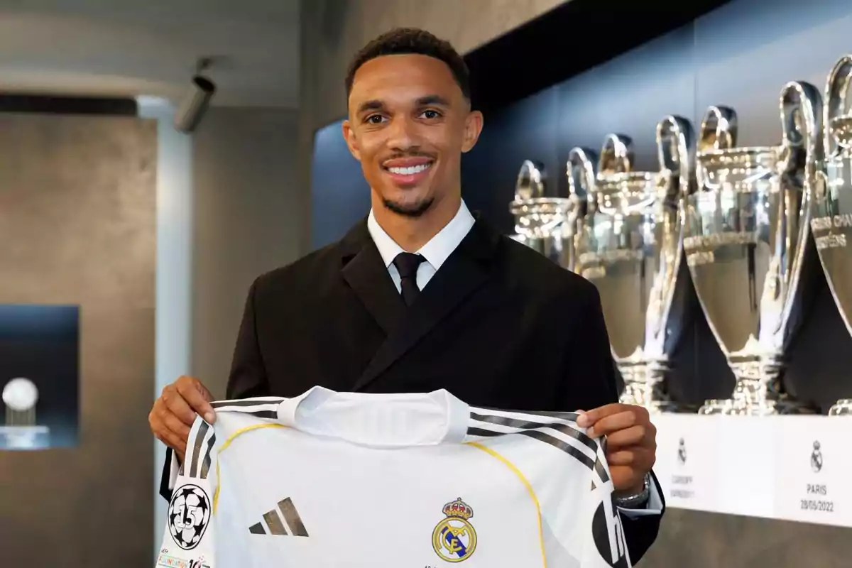 A smiling man in a suit holds a Real Madrid jersey in front of several Champions League trophies. A smiling man in a suit holds a Real Madrid jersey in front of several Champions League trophies.