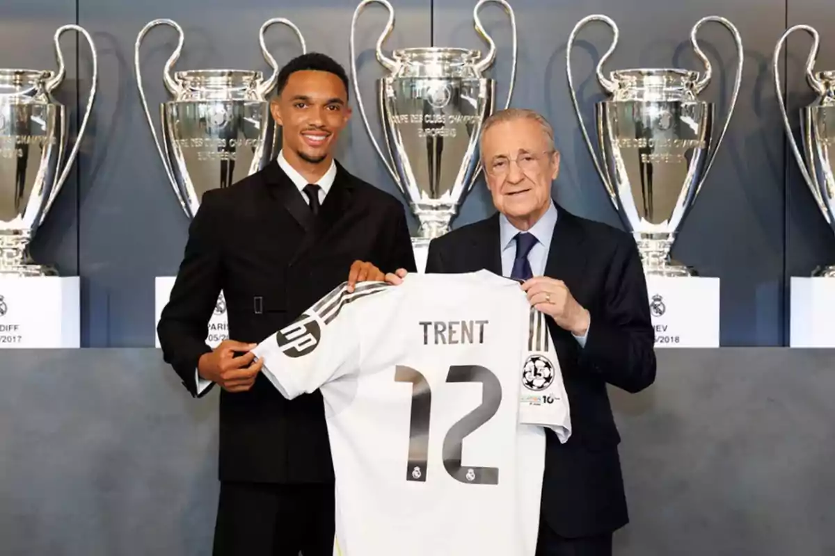 Two men pose smiling, holding a white soccer jersey with the name Trent and the number 12 in front of several large Champions League trophies. Two men pose smiling, holding a white soccer jersey with the name Trent and the number 12 in front of several large Champions League trophies.