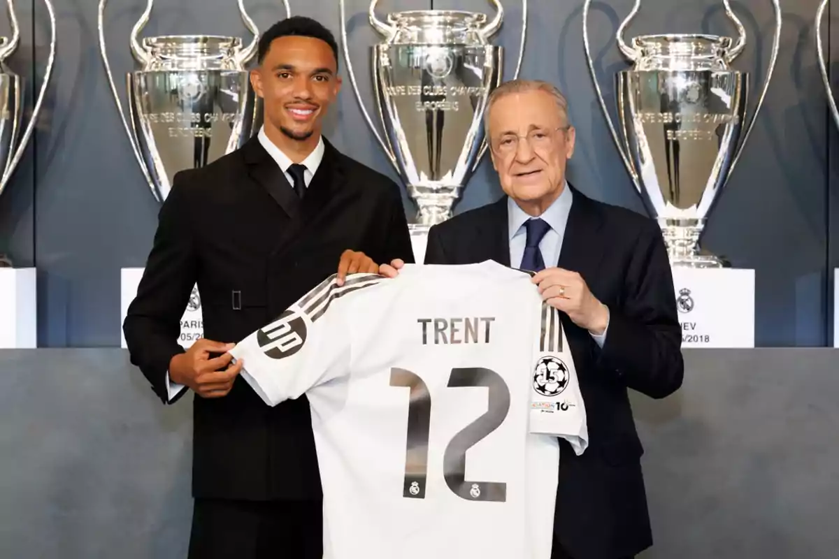 Two men pose smiling, holding a white jersey with the name Trent and the number 12 in front of several silver soccer trophies. Two men pose smiling, holding a white jersey with the name Trent and the number 12 in front of several silver soccer trophies.