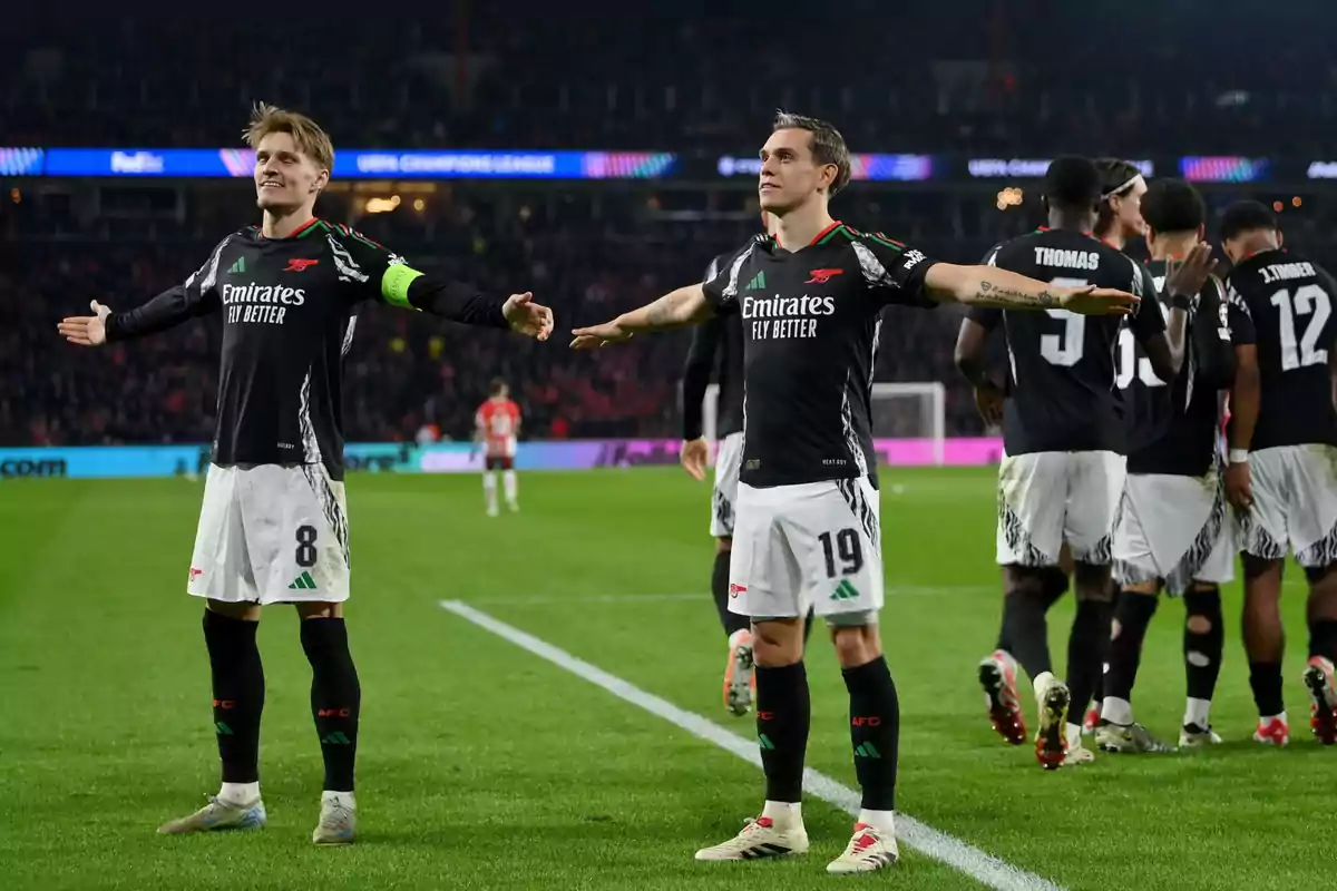 Soccer players celebrating on the field with arms outstretched, wearing black and white uniforms, with other players in the background.