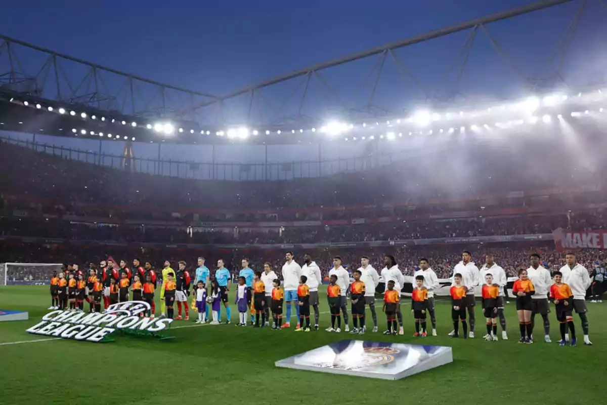 Jugadores de dos equipos de fútbol alineados en el campo antes de un partido de la Champions League, con niños acompañándolos y un estadio lleno de espectadores iluminado por potentes luces. Jugadores de dos equipos de fútbol alineados en el campo antes de un partido de la Champions League, con niños acompañándolos y un estadio lleno de espectadores iluminado por potentes luces.