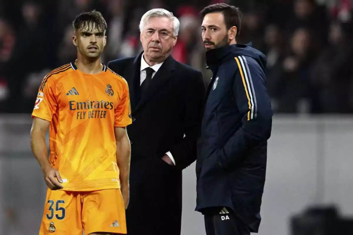 Three men on a soccer field, one of them wearing an orange Real Madrid uniform and the other two wearing warm clothes, appear to be having a conversation.