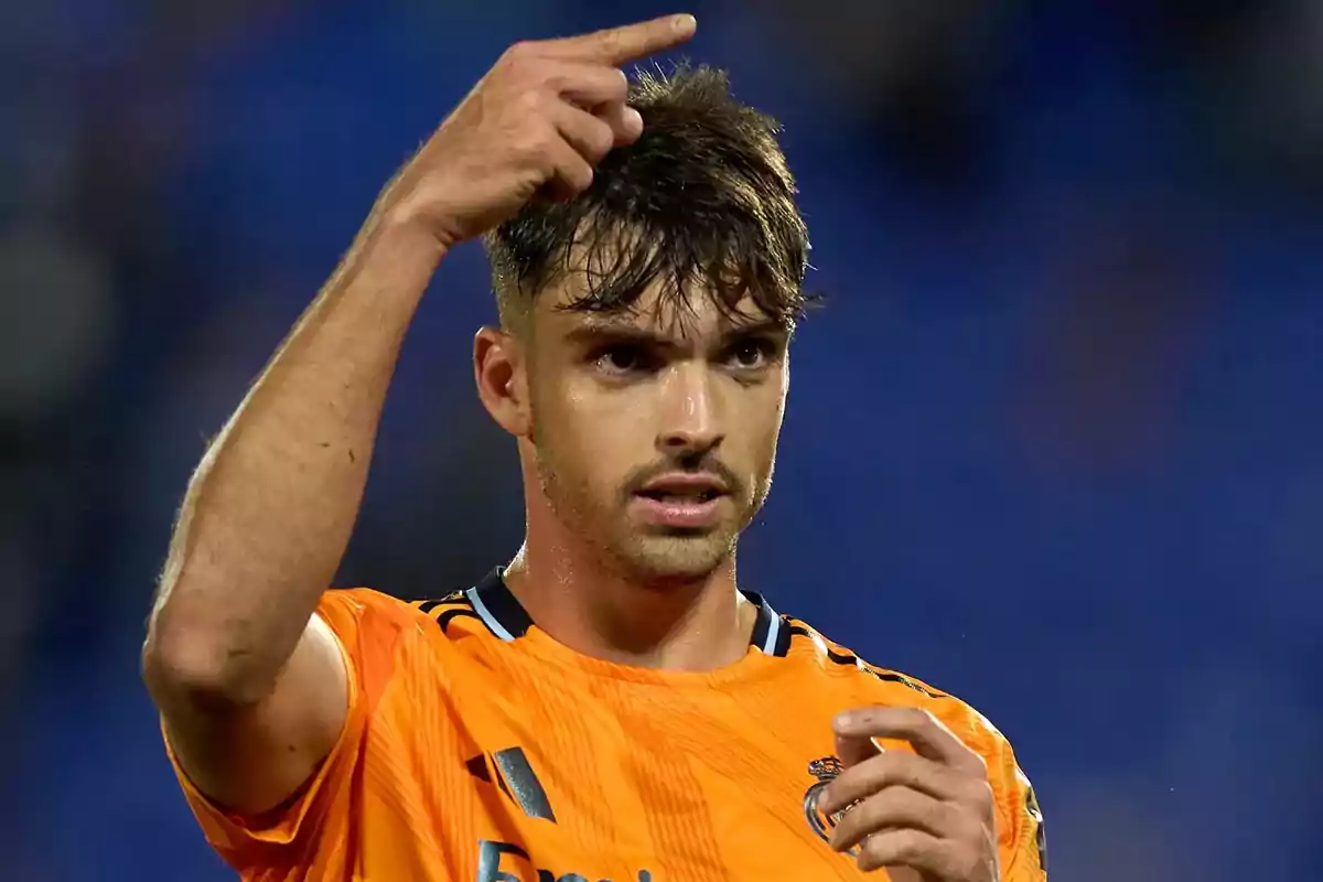 Un jugador de fútbol con camiseta naranja señala hacia arriba con el dedo en un estadio.