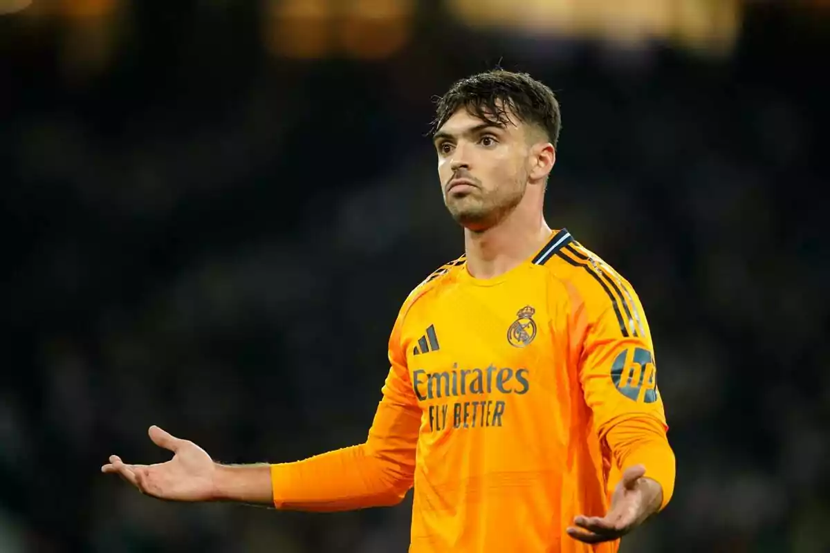 Un jugador de fútbol con uniforme naranja del Real Madrid en el campo con los brazos extendidos.