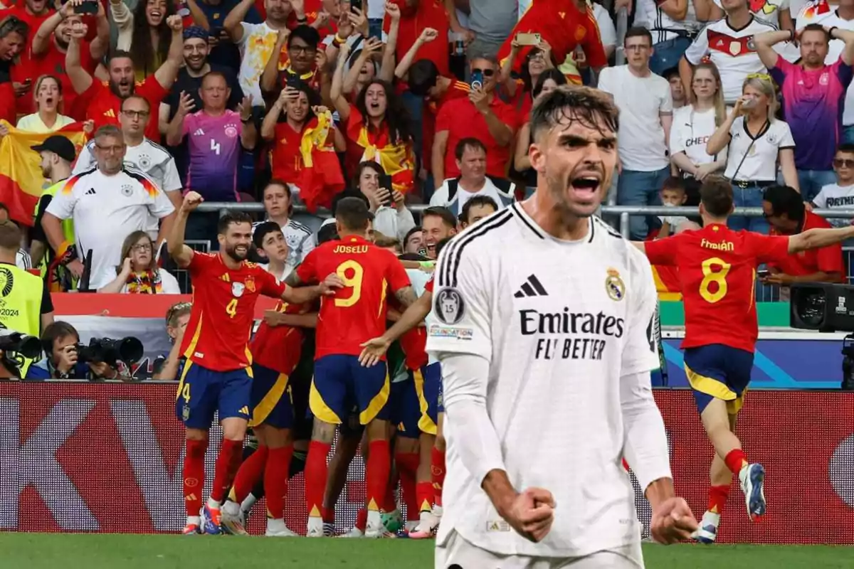 Un jugador de fútbol con uniforme blanco celebra con el puño cerrado mientras al fondo un grupo de jugadores con uniforme rojo se abrazan frente a una multitud de aficionados.