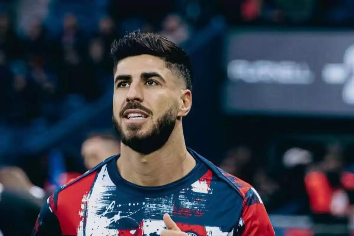 Un jugador de fútbol con barba y cabello oscuro lleva una camiseta deportiva colorida mientras está en un estadio.