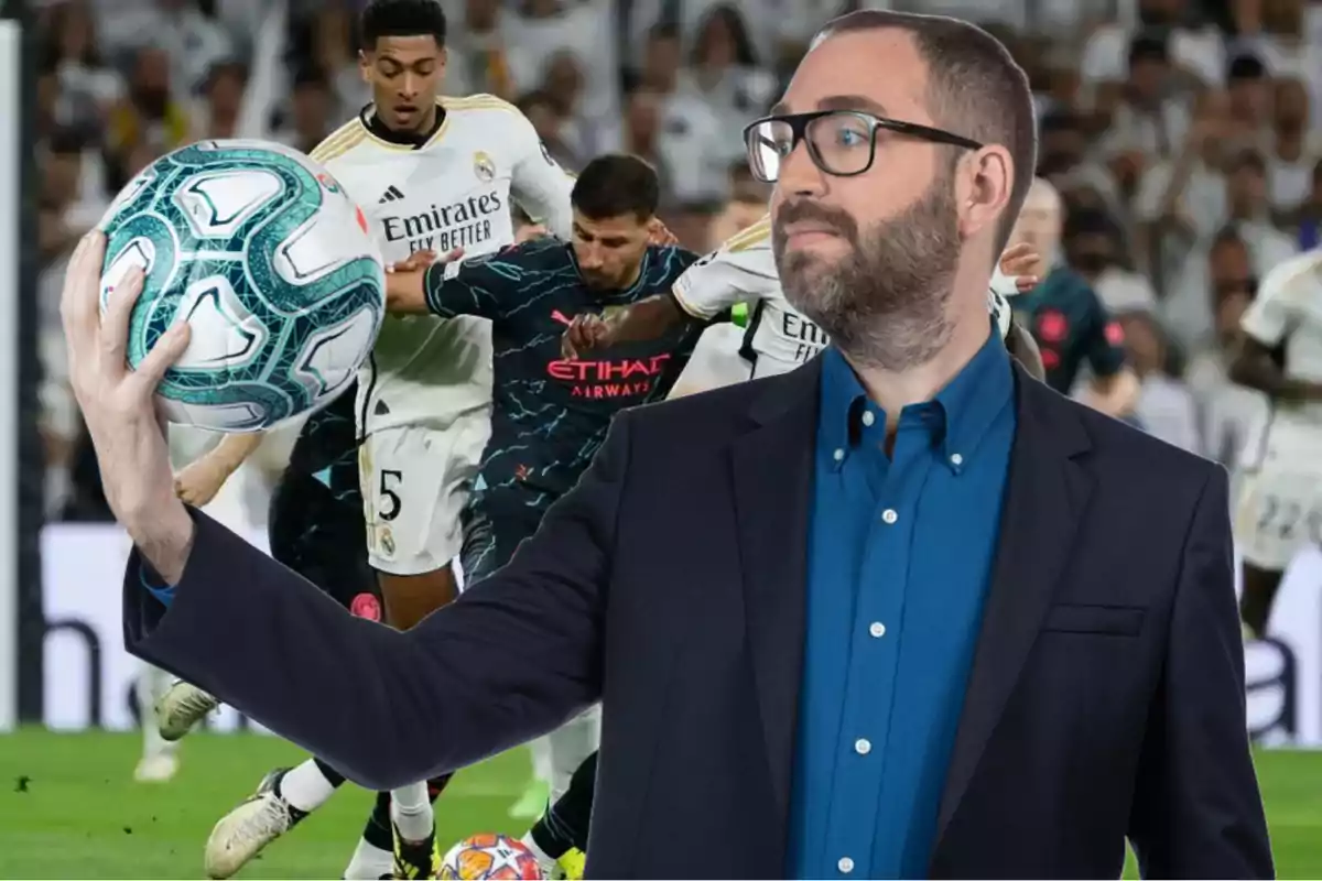 Un hombre con gafas y barba sostiene un balón de fútbol mientras en el fondo se desarrolla un partido entre jugadores con uniformes blancos y oscuros.