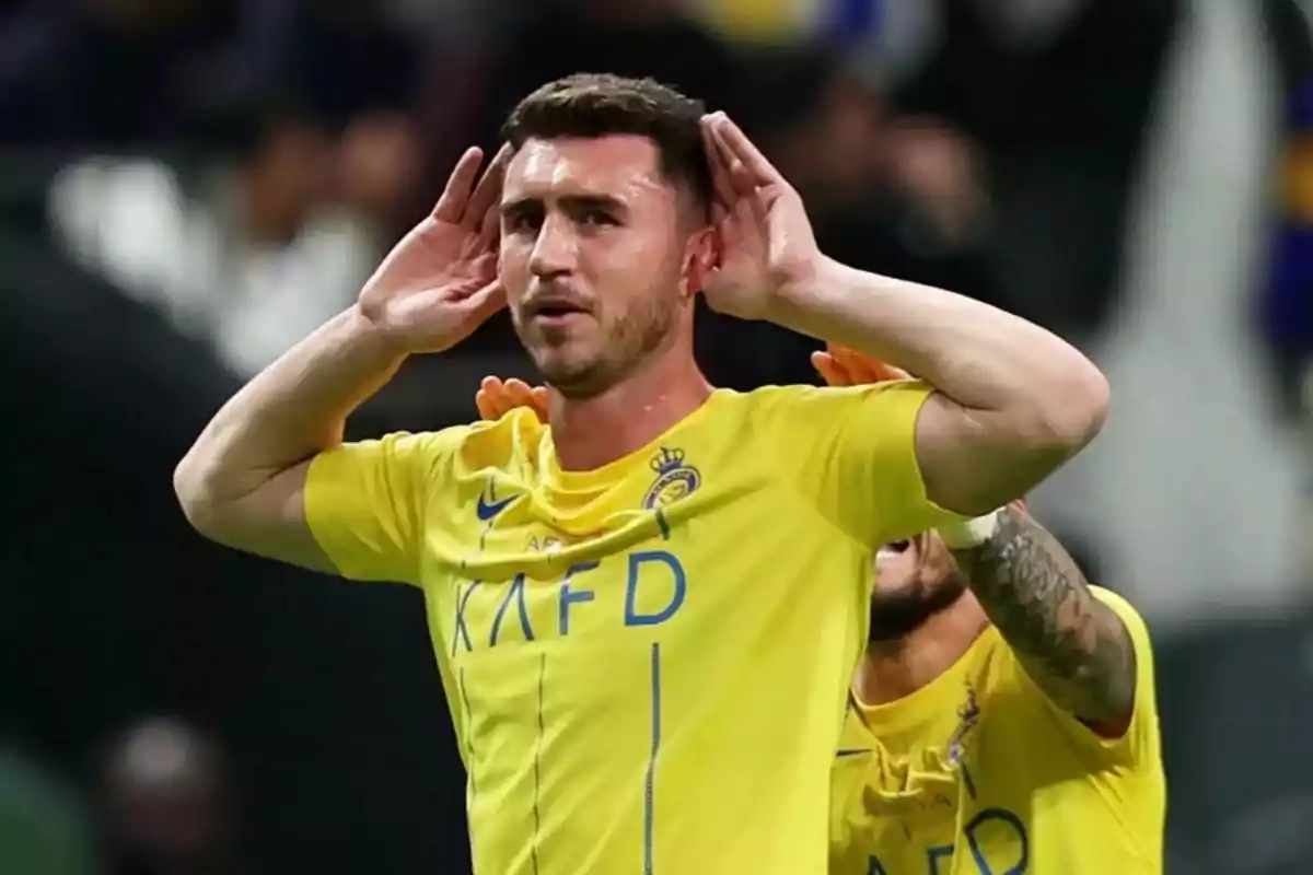 Jugador de fútbol celebrando un gol con las manos en las orejas mientras viste un uniforme amarillo en un estadio.