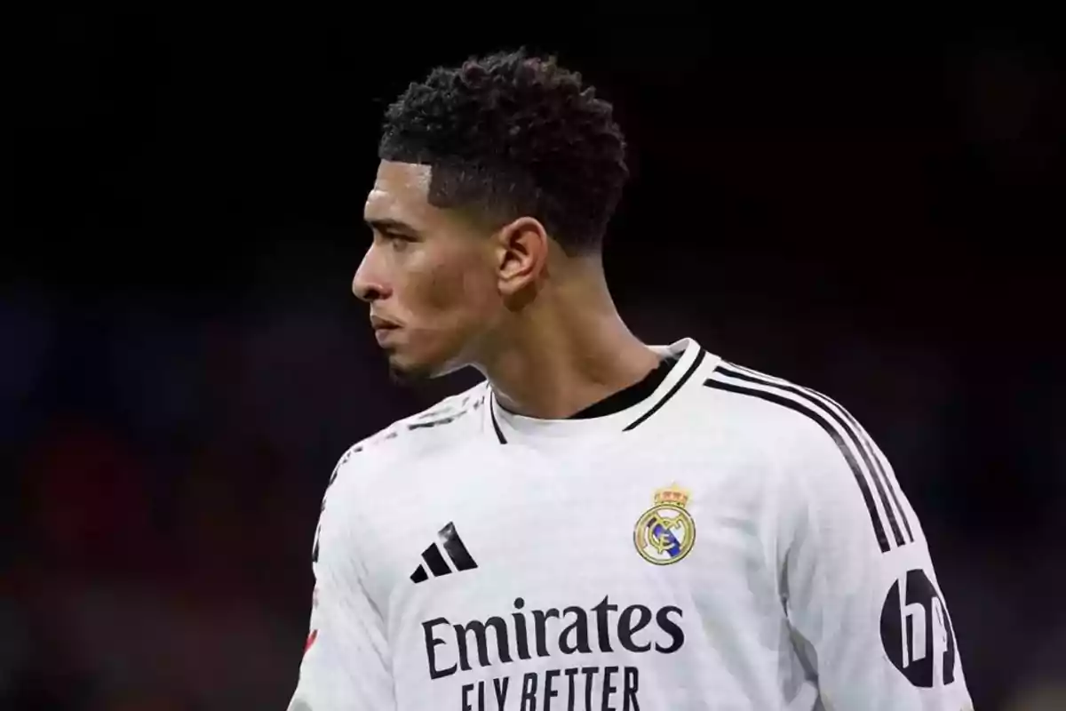 Jugador de fútbol con uniforme blanco del Real Madrid mirando hacia la izquierda en un fondo oscuro.