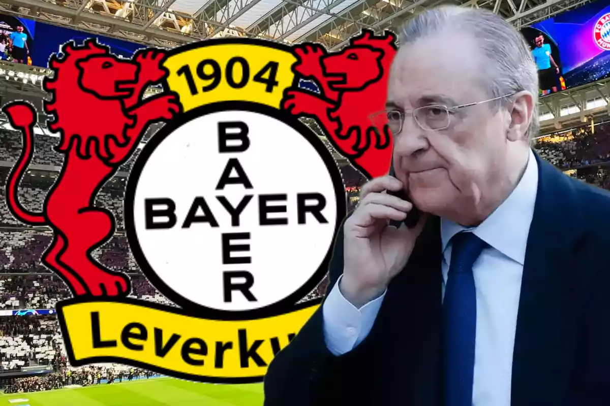 An older man with glasses and a dark suit talks on the phone in front of the Bayer Leverkusen logo at a soccer stadium.