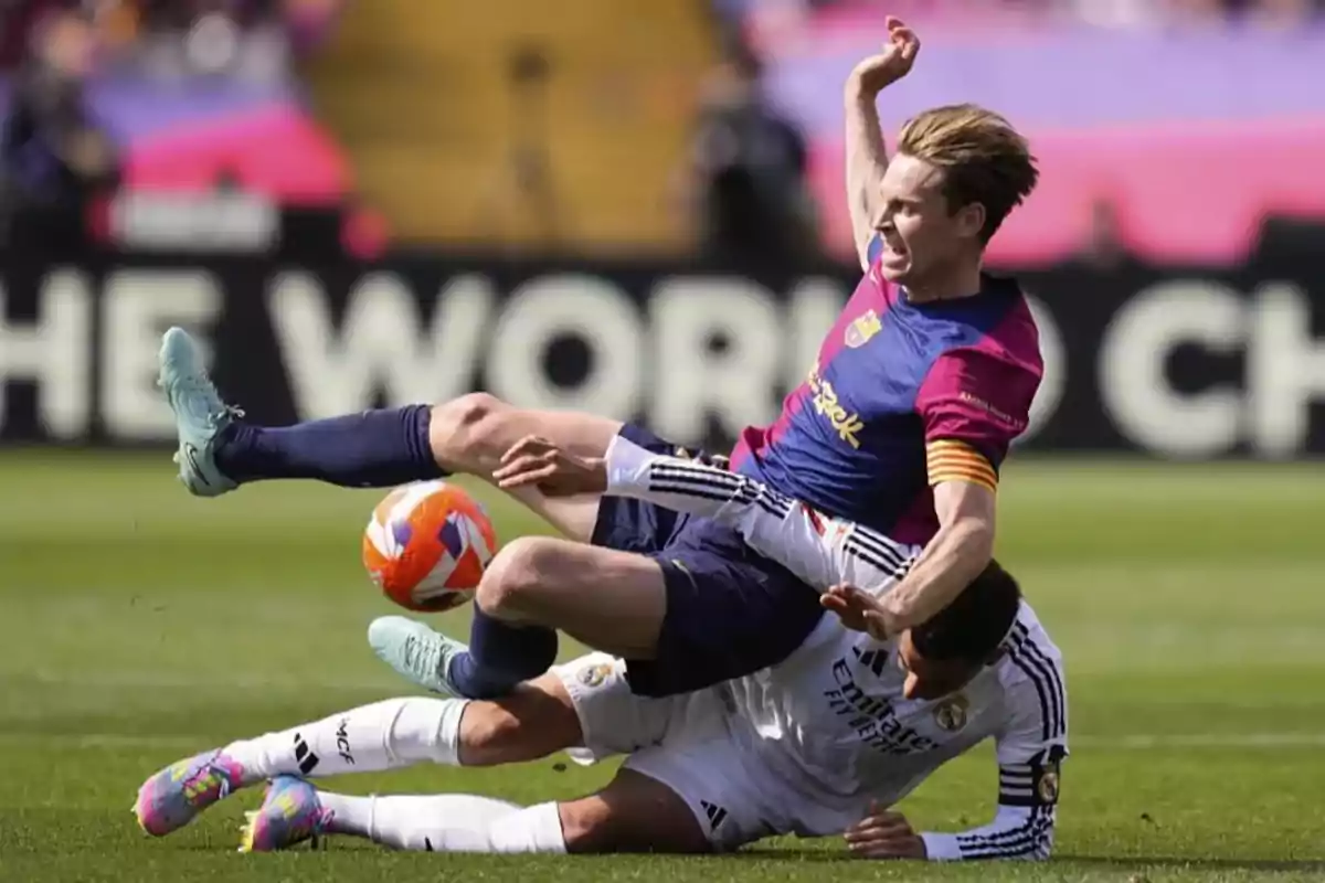 Two players compete for the ball on the field, one of them is on the ground while the other jumps over him.