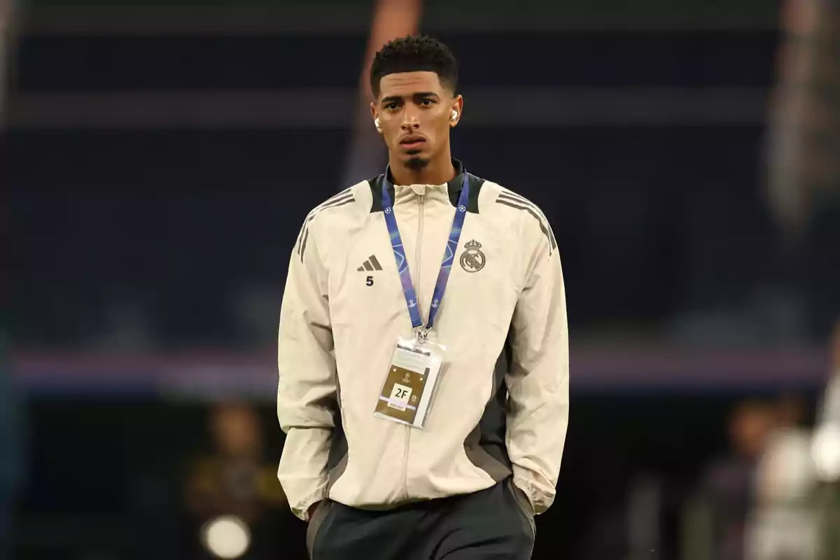 Un jugador de fútbol con una chaqueta del Real Madrid y un gafete colgado del cuello está de pie en un campo deportivo. Un jugador de fútbol con una chaqueta del Real Madrid y un gafete colgado del cuello está de pie en un campo deportivo.