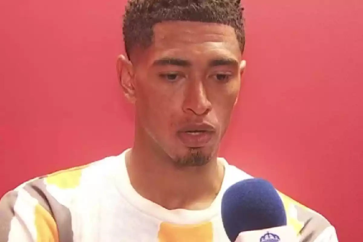 A man with a serious expression speaks in front of a microphone with a red background.